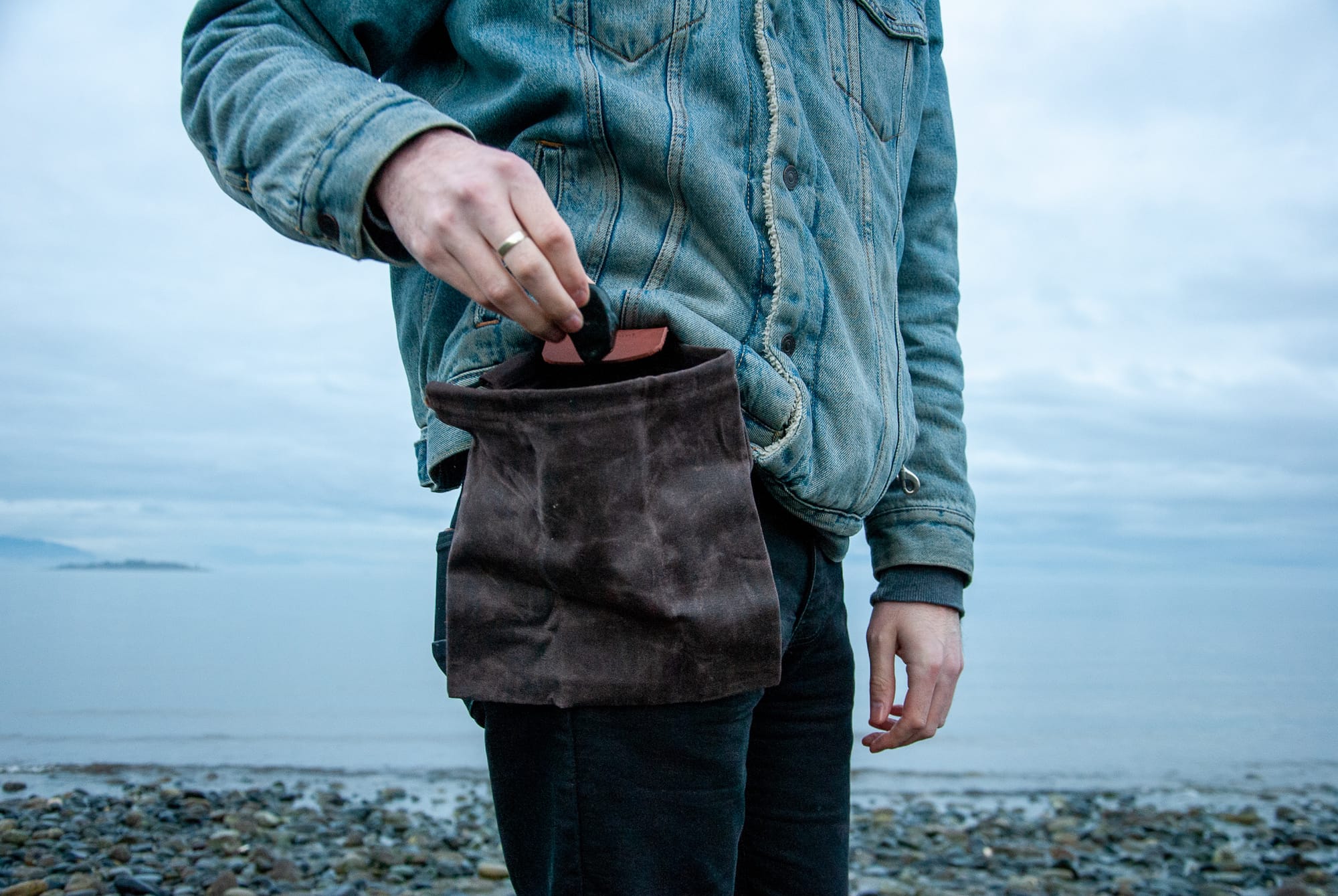 Man in a light denim jacket places a rock in his brown canvas foraging pouch while standing on the beach