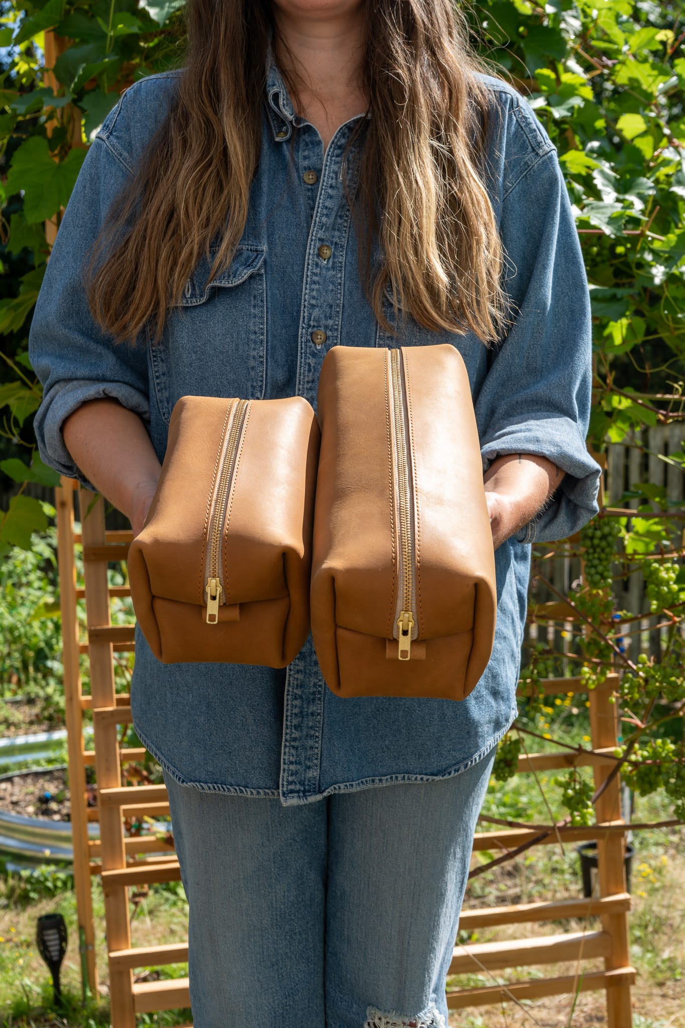a person in a denim shirt and jeans holding two tan toiletry bags to show their zippers