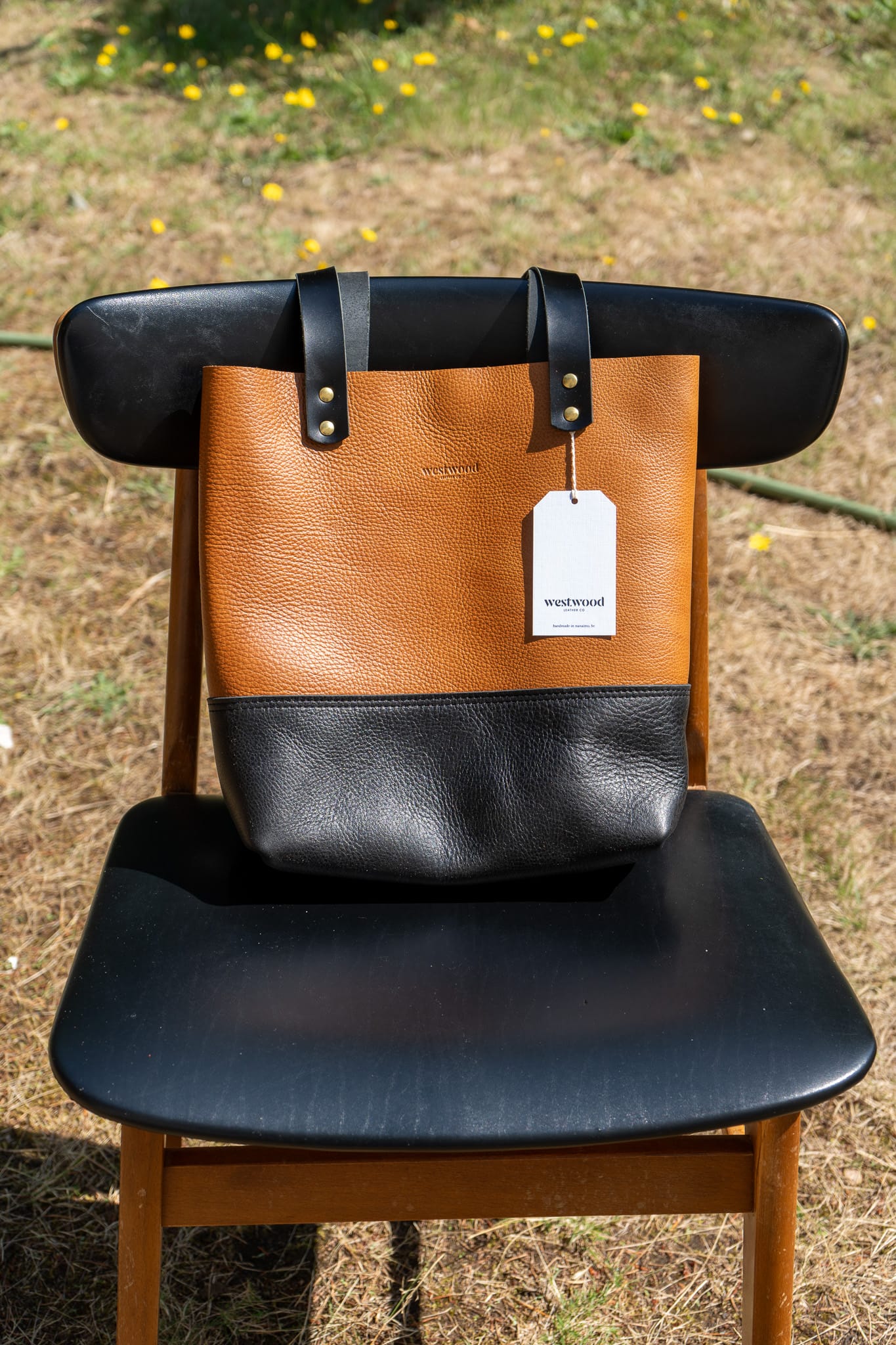 Brown and black leather bag with a tag on a wooden chair outdoors.