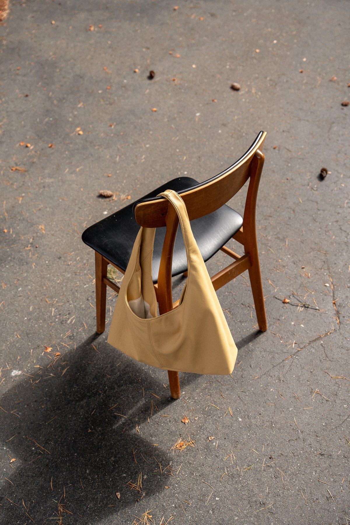 A yellow leather tote bag hangs from the back of a wooden chair with a black seat, placed on an asphalt ground outdoors.