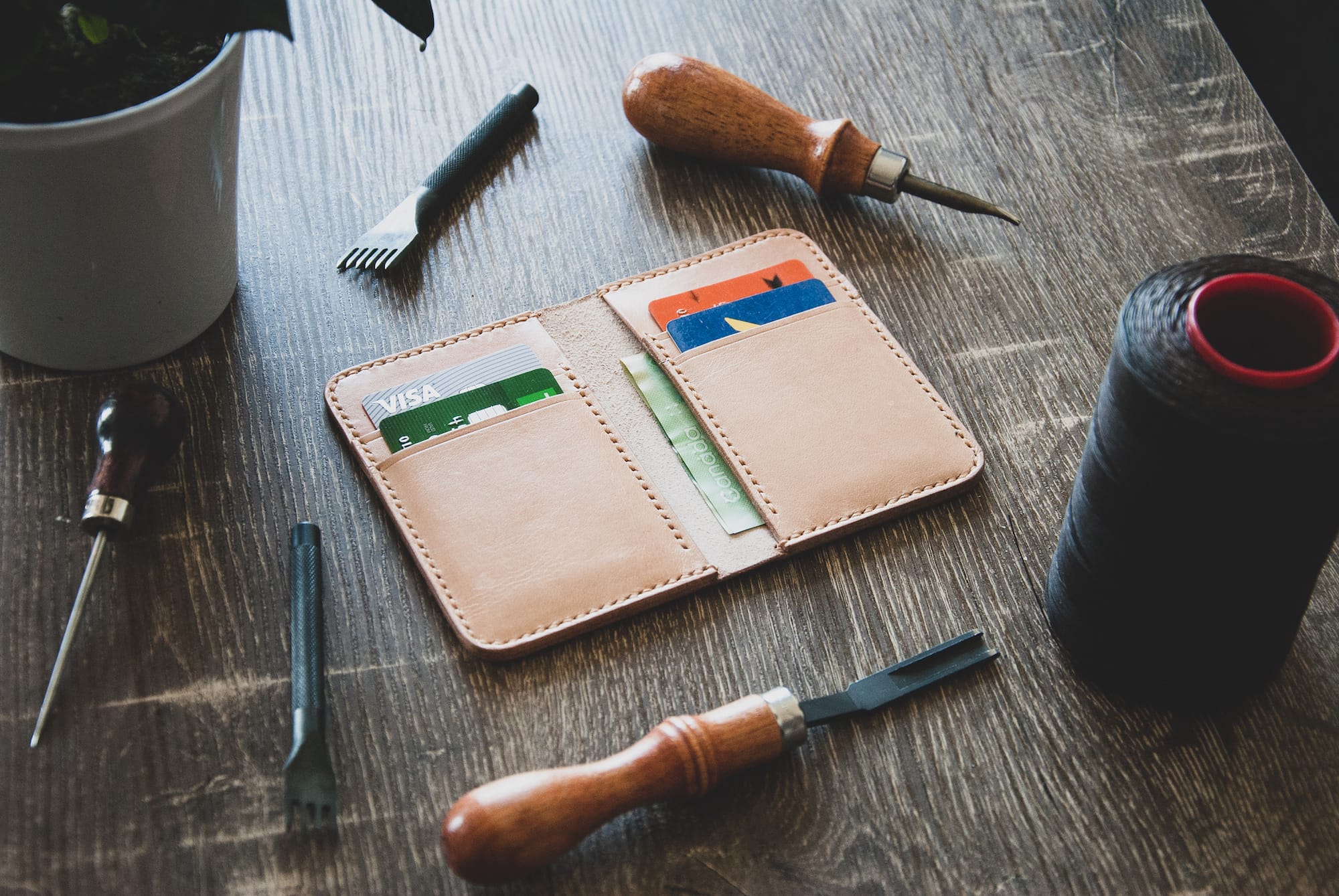 leather wallet with credit cards and cash on a wooden table with leatherworking tools and a spool of thread