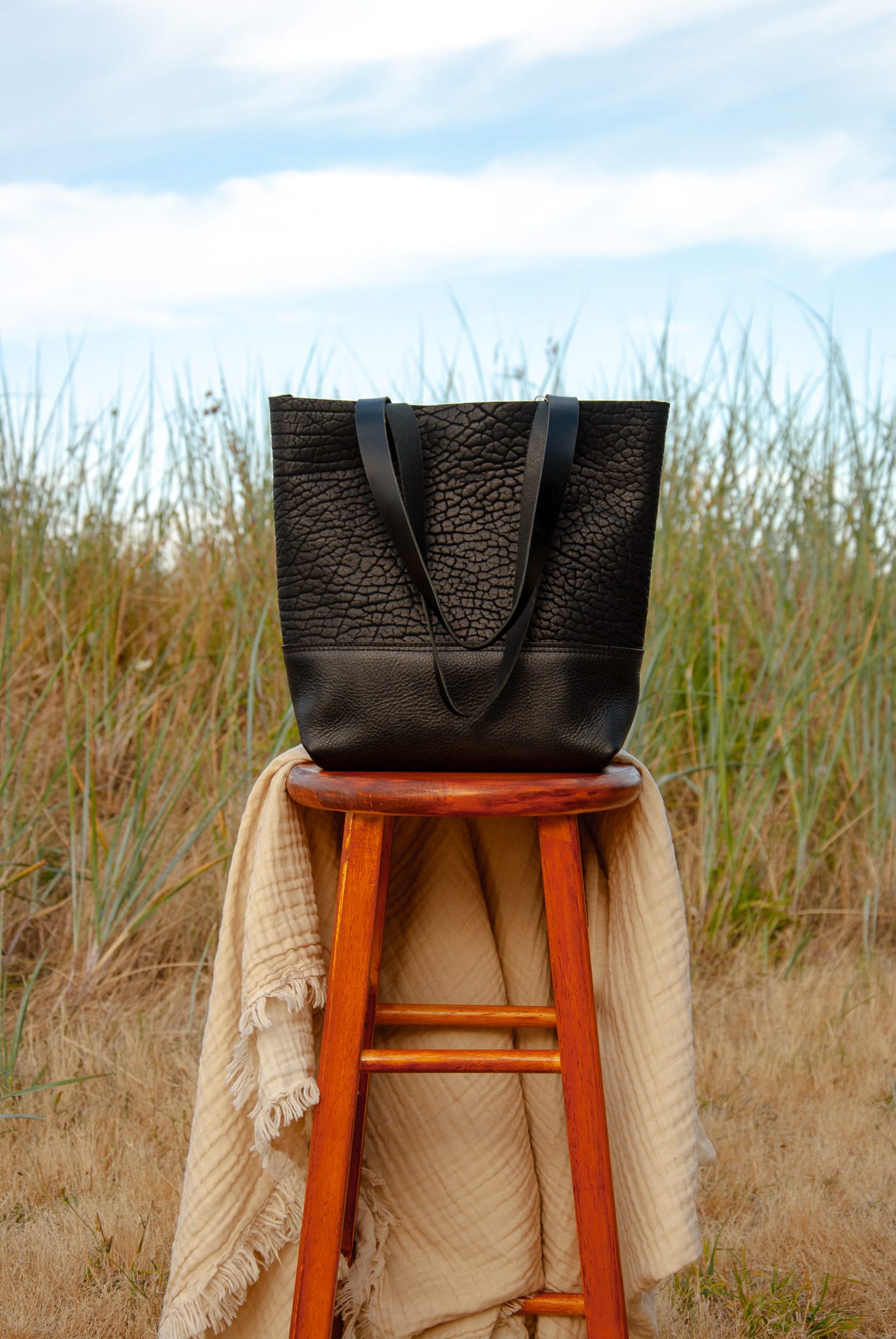 Black leather tote bag on a wooden stool with a beige blanket in a grassy outdoor setting