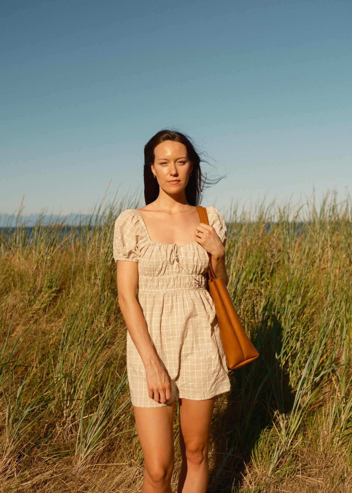 Woman in a beige dress holding a brown bag in a grassy field with a clear blue sky.