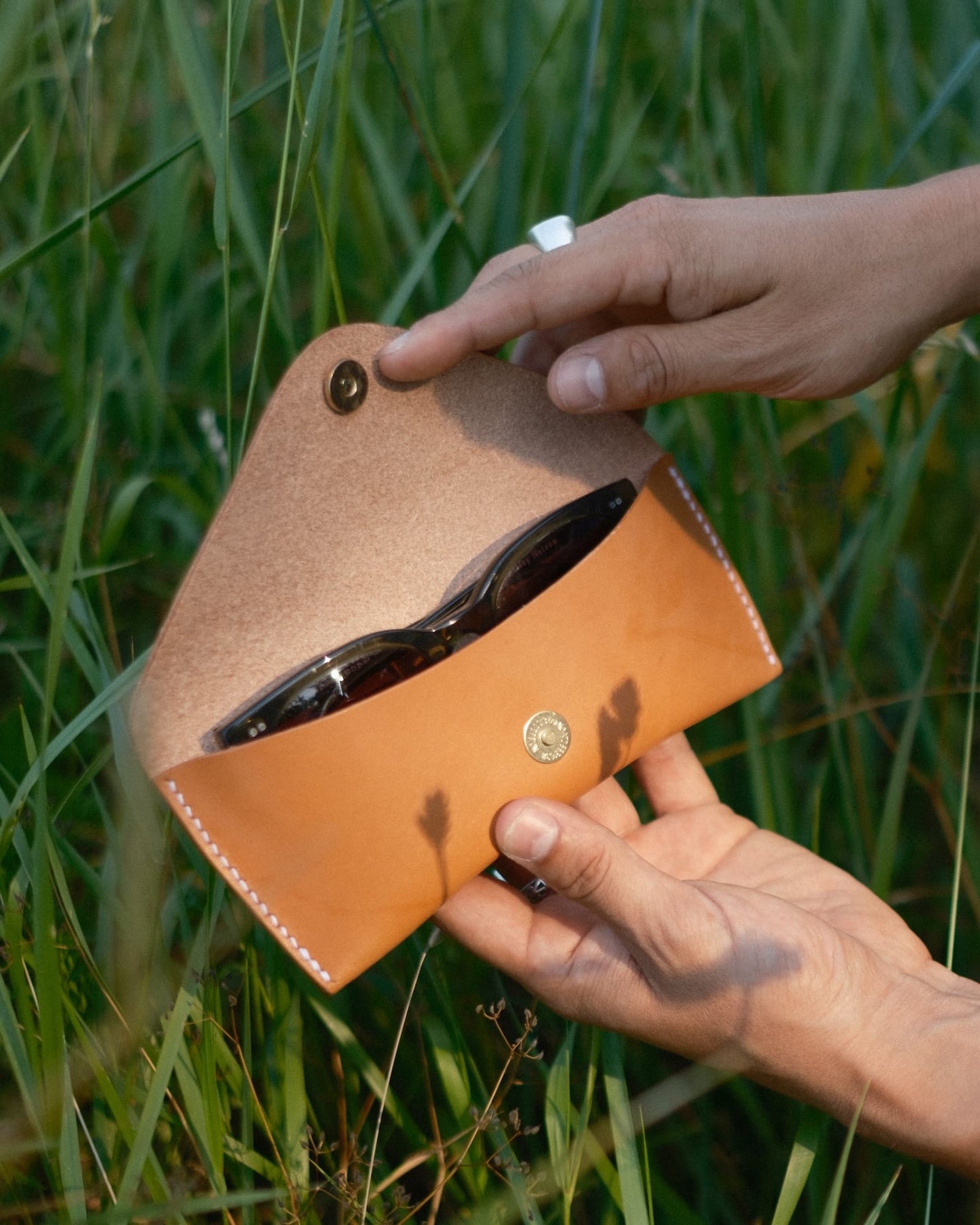Person holding a brown leather case with sunglasses against a grassy background