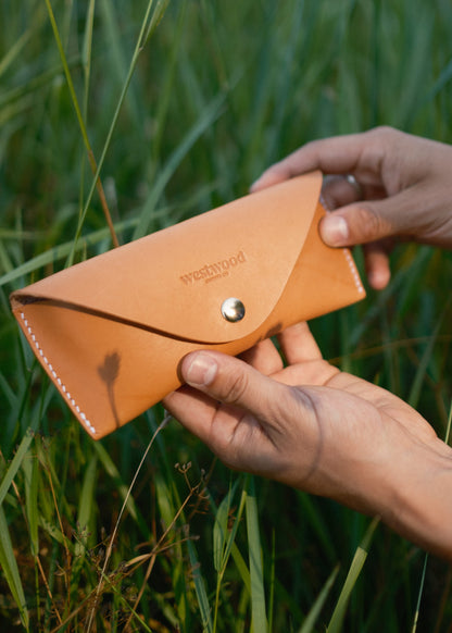 Person holding a brown leather glasses case with 'Westwood' branding in a grassy outdoor setting