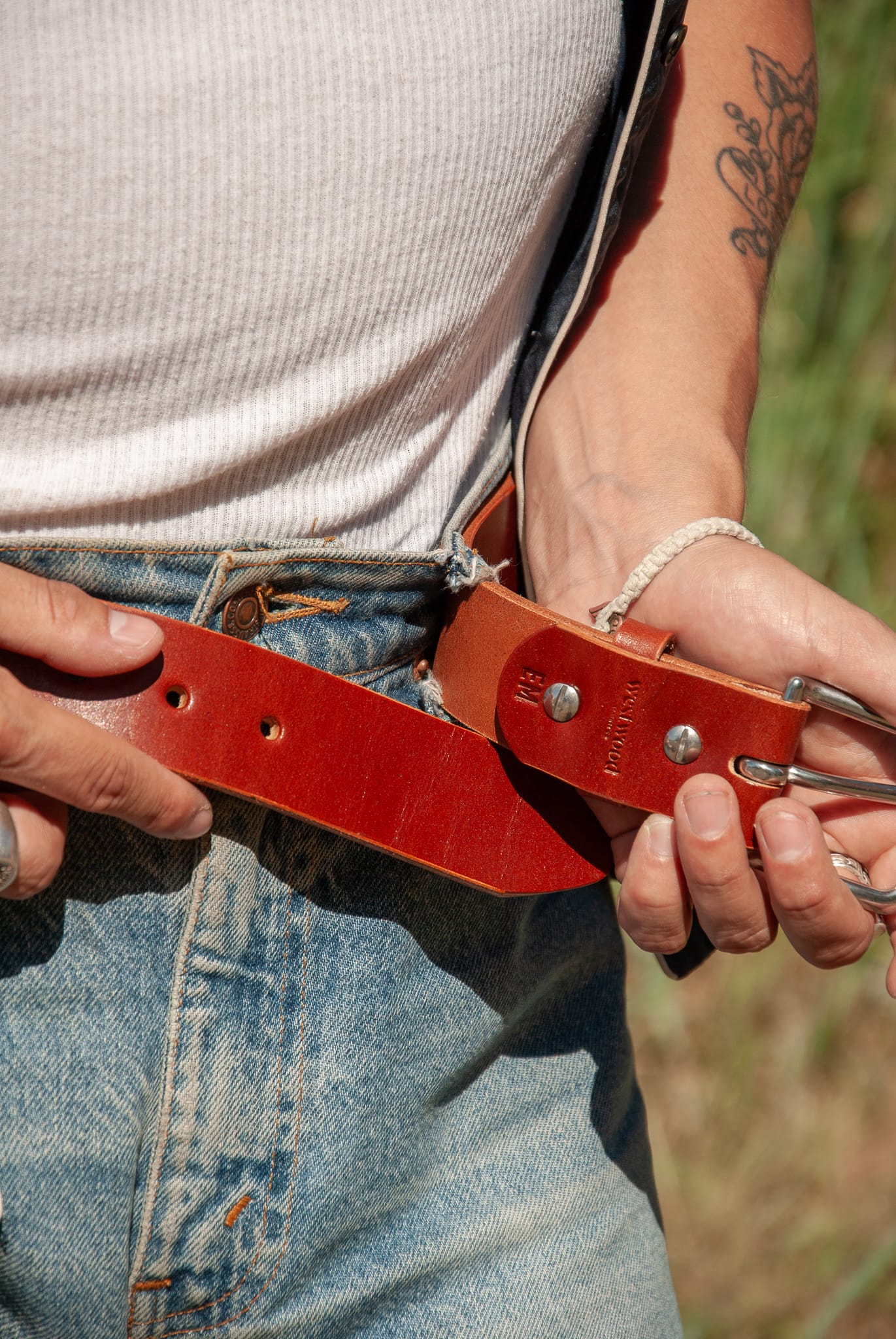 Person adjusting a red leather belt with a buckle in a natural setting.