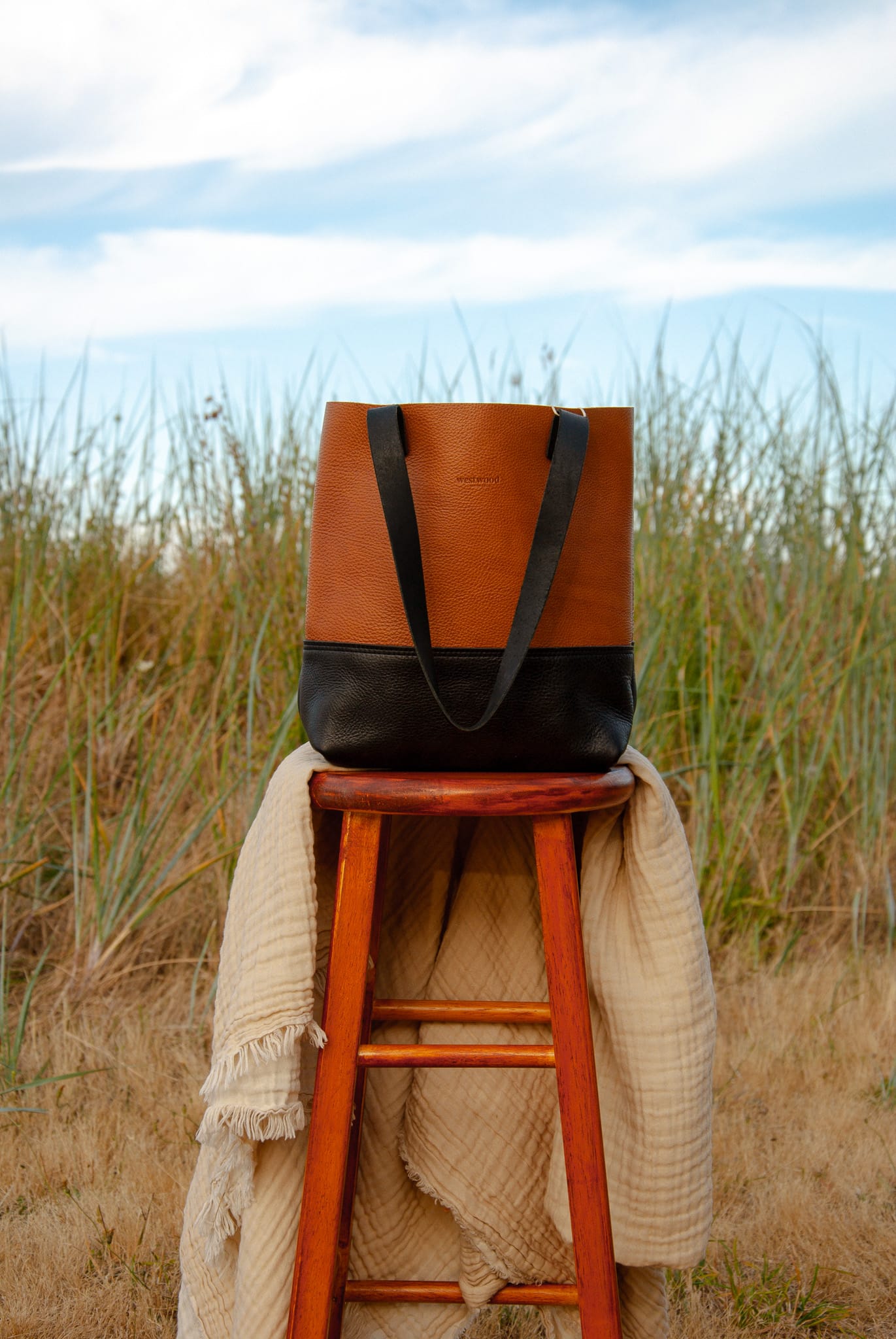 Brown and black tote bag on a wooden stool with a blanket in a grassy field