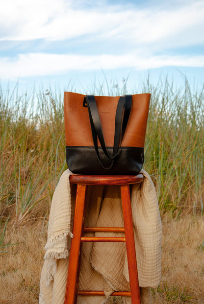 Brown and black tote bag on a wooden stool with a beige blanket in a grassy field.