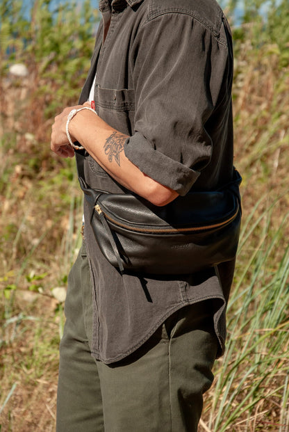 Person wearing a dark shirt and green pants with a black bag over one shoulder, standing in a natural setting.