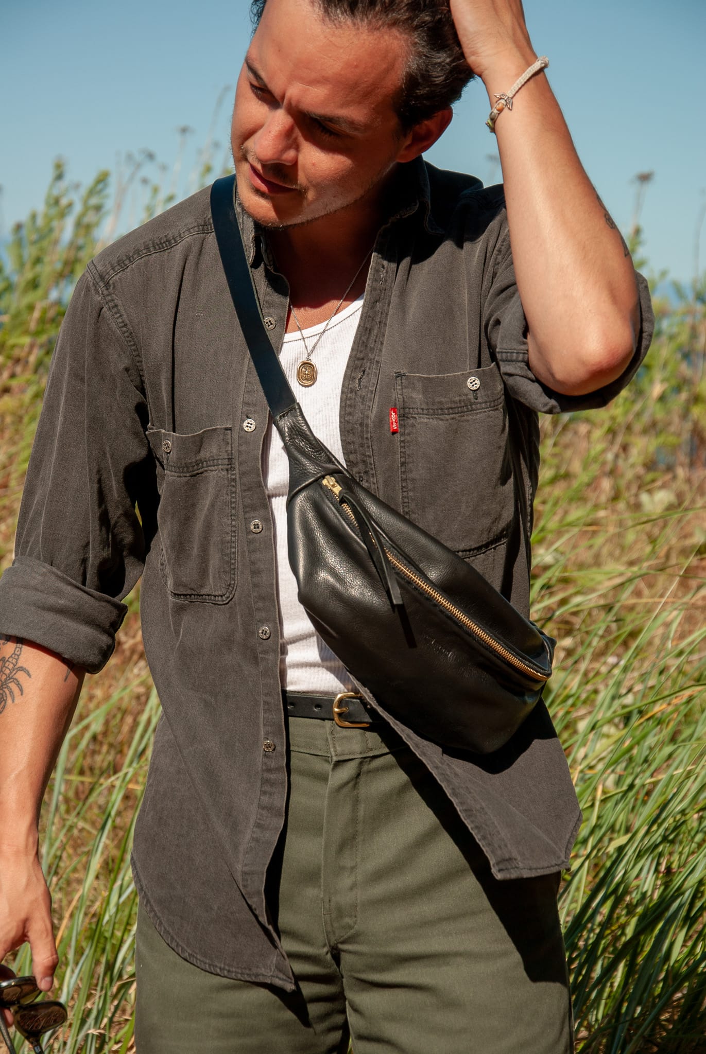 Man wearing a dark shirt and green pants with a black bag over his shoulder in a grassy field.