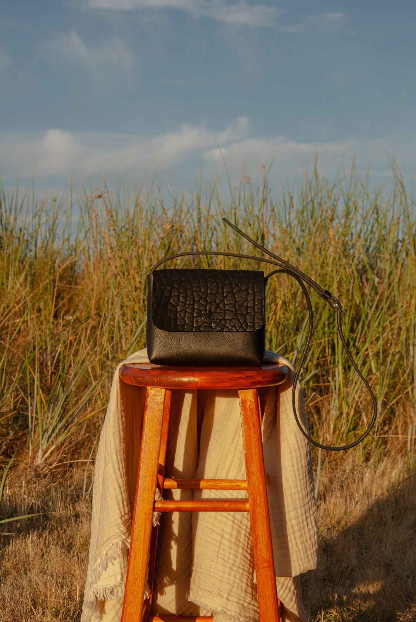 Black handbag on a wooden stool with tall grass and blue sky in the background