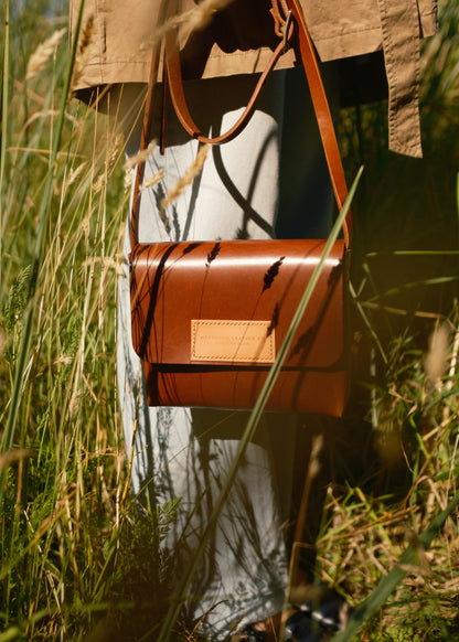 Brown leather bag hanging from a person's hand in a grassy field