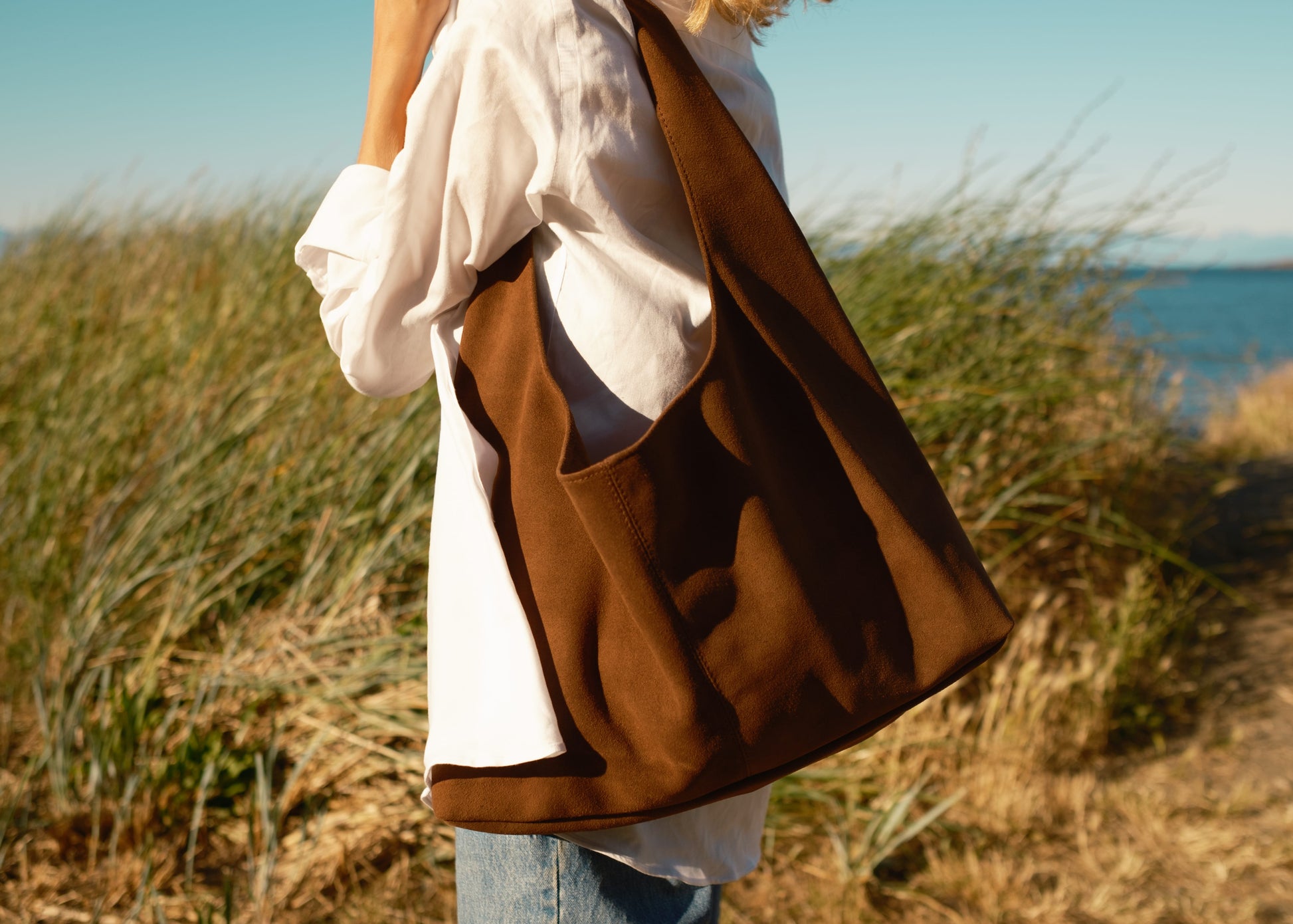 Person holding a brown suede bag on a grassy hillside with ocean view
