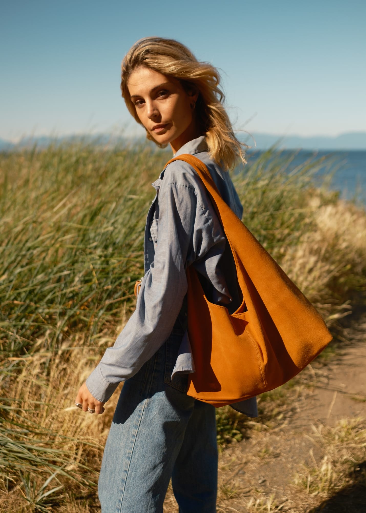 Woman with an orange tote bag walking through a grassy field with a scenic background