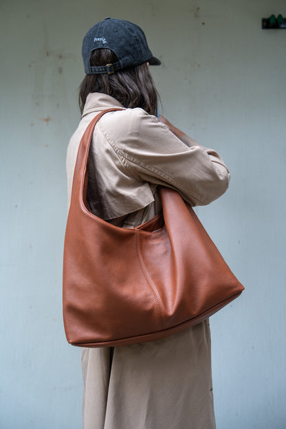 A person shown from the back wearing a dark blue baseball cap, a beige trench coat, and carrying a large brown leather hobo shoulder bag against a light blue-gray wall.
