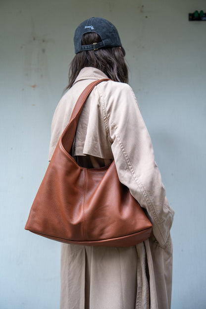 A person shown from behind wearing a beige trench coat and a dark blue baseball cap, carrying a large brown leather hobo shoulder bag against a light blue-gray wall.