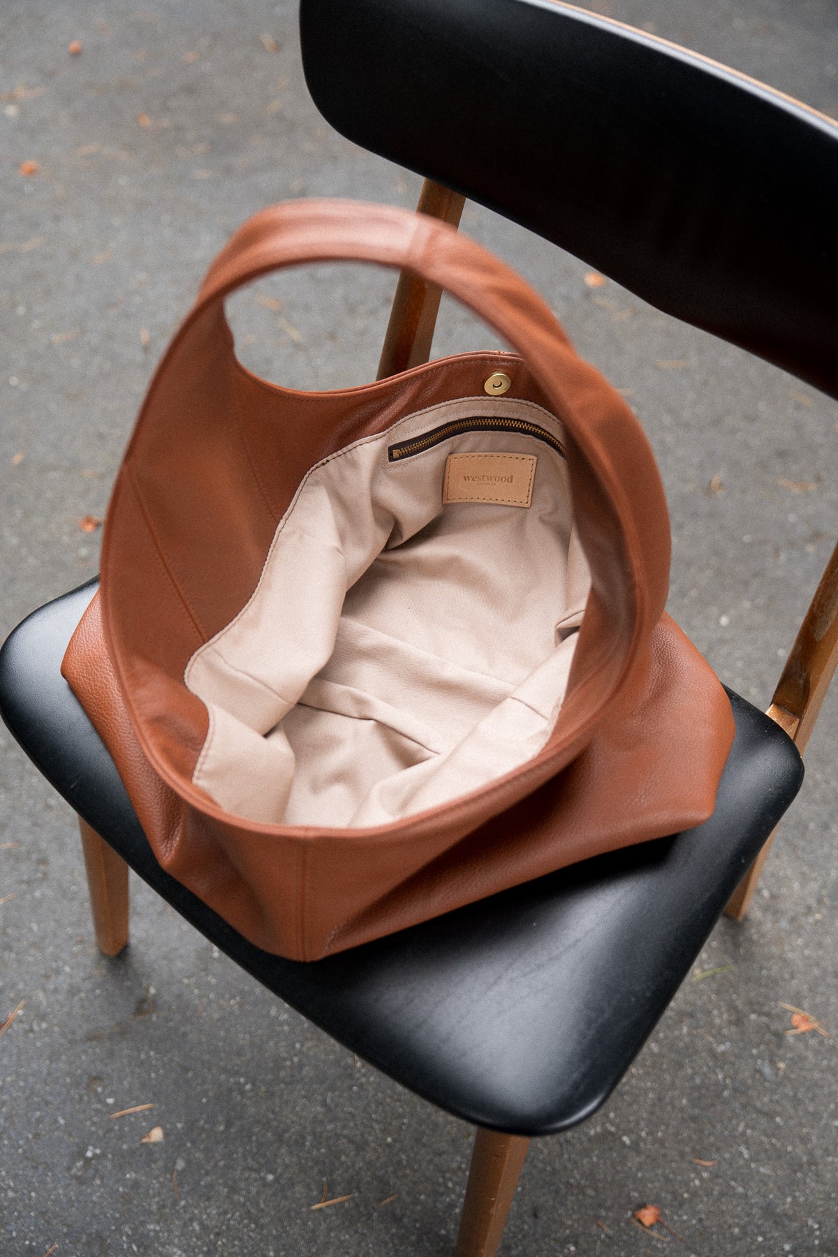 A tan leather hobo shoulder bag with a beige interior lining, resting on a black leather chair with wooden legs on an asphalt ground.