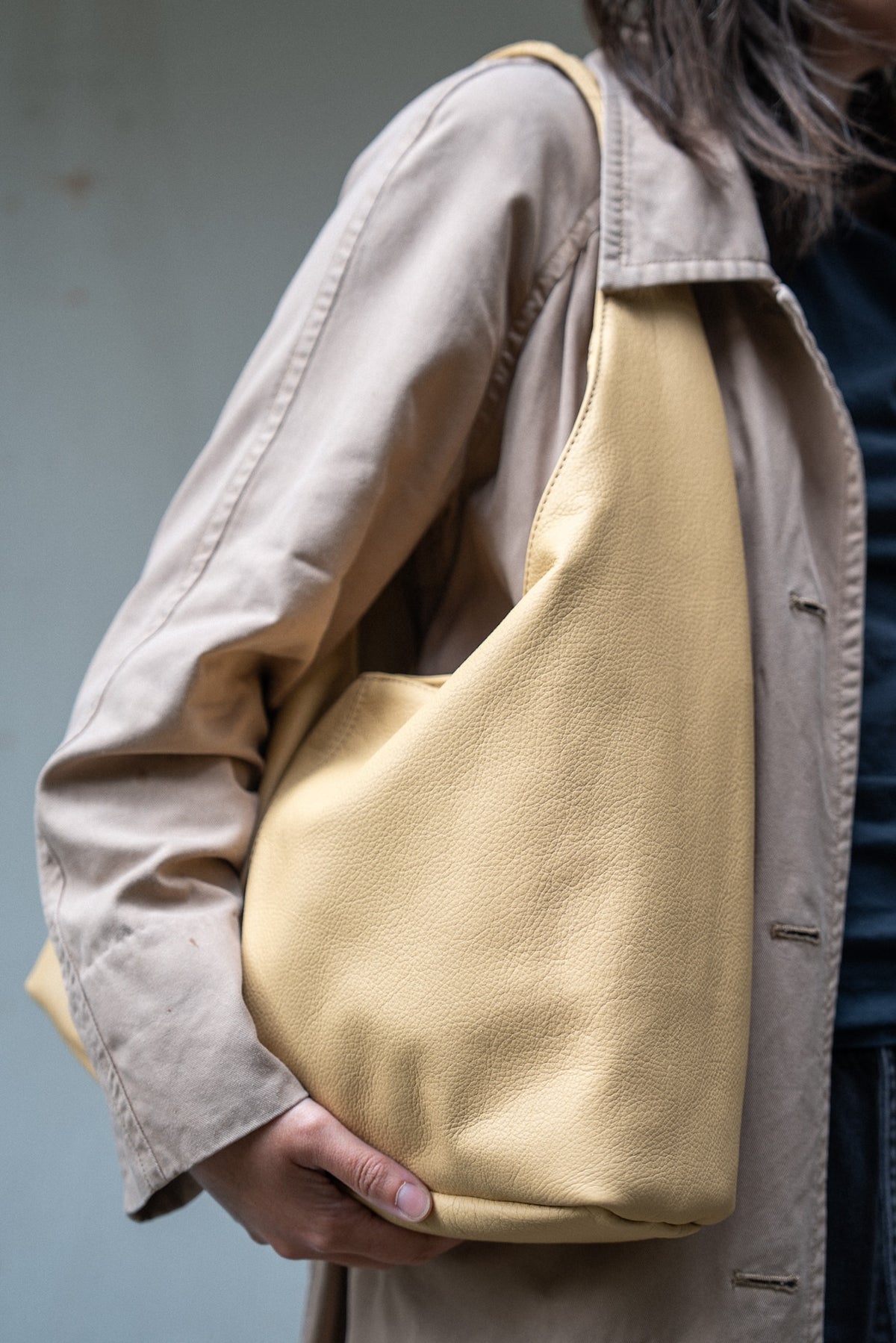 A person wearing a beige trench coat carries a large, soft slouchy shoulder bag in a pale yellow-beige pebbled leather. The person is also wearing a dark blue shirt.