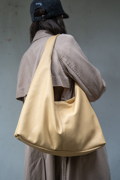 A person shown from behind wearing a beige trench coat and a dark cap, carrying a large, soft slouchy shoulder bag in a light beige or cream leather. The background is a textured, muted gray wall.