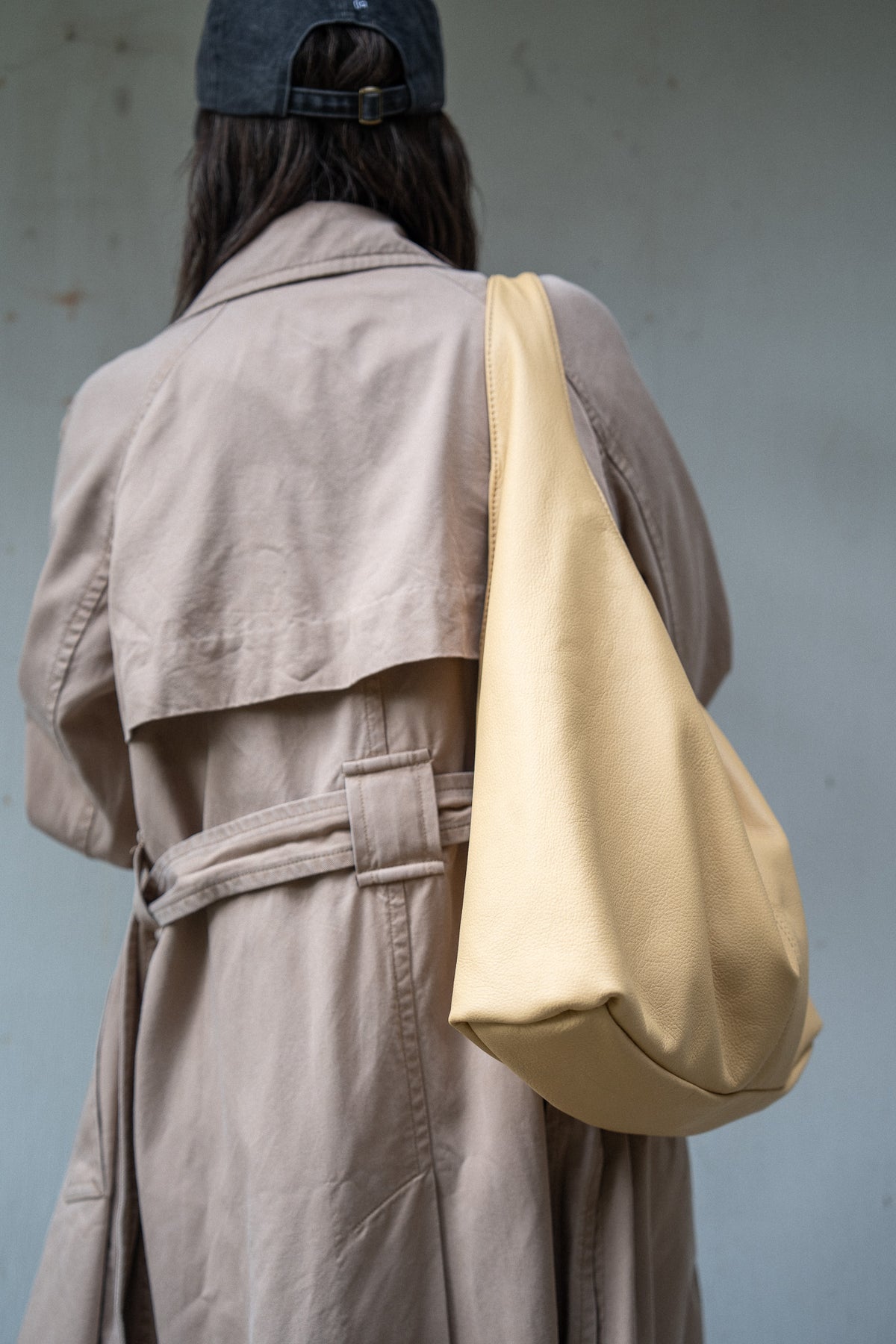 A person viewed from behind wearing a beige trench coat and a dark cap, carrying a large soft slouchy shoulder bag in a pale yellow-beige color against a muted gray wall.