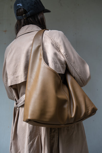 A person shown from behind wearing a beige trench coat and a dark cap, carrying a large tan leather shoulder bag against a muted blue-gray wall.