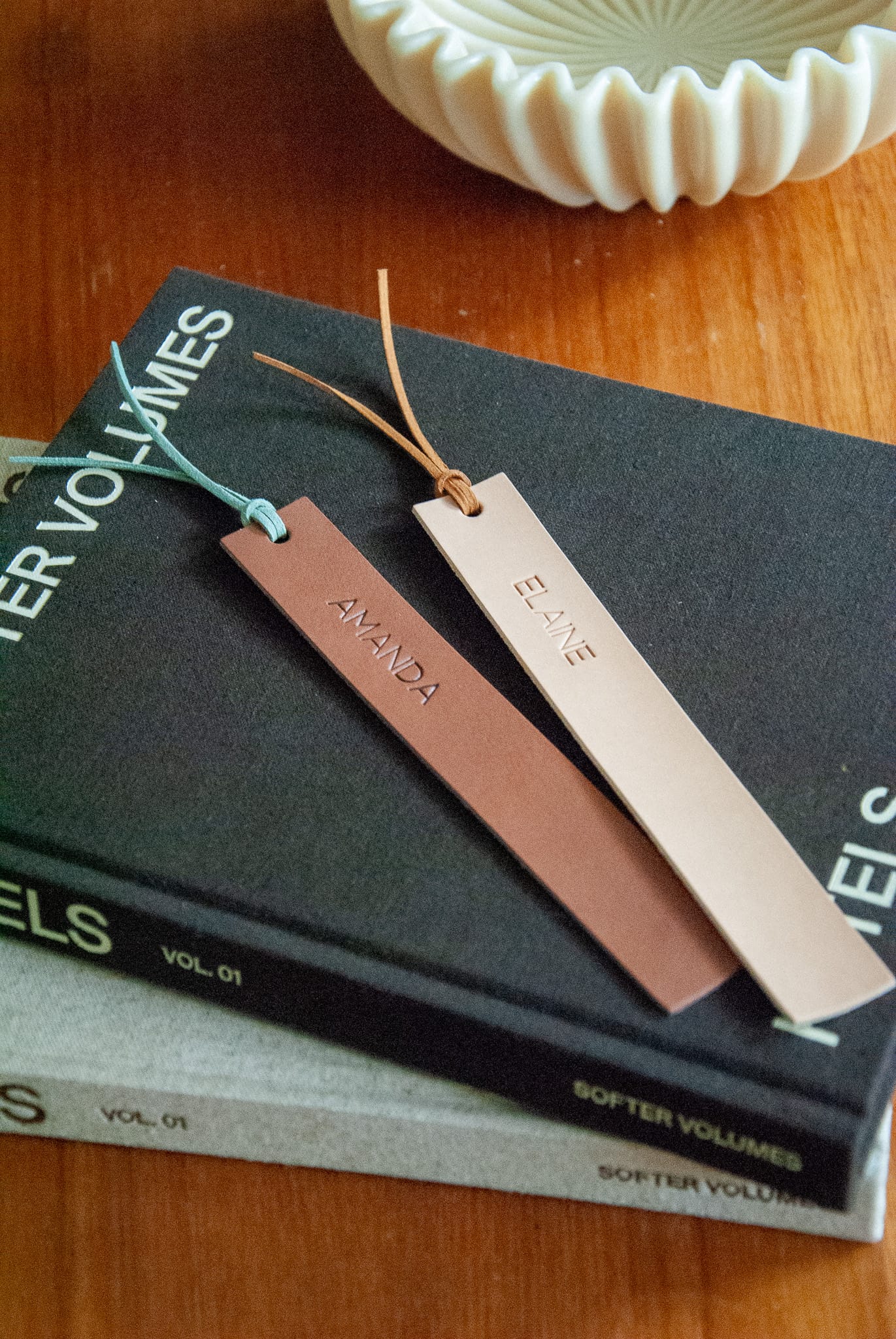 Two leather bookmarks on a stack of books with a decorative bowl in the background.