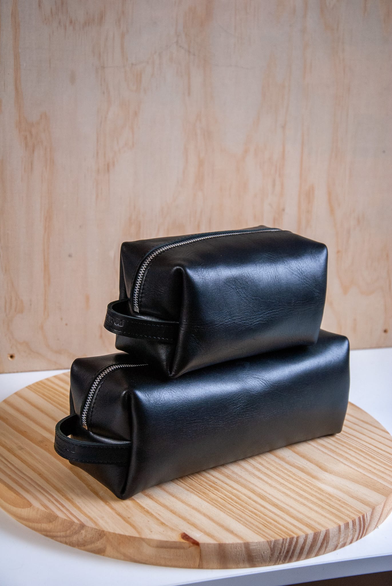 Two black leather toiletry bags stacked on a wooden surface with a light wood panel background.